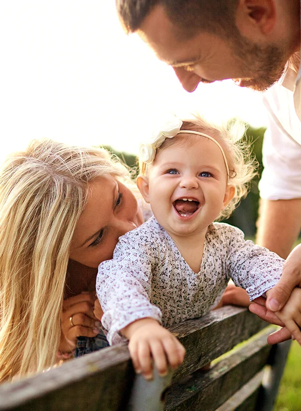 A baby stands on a bench, smiling surrounded by her mother and father